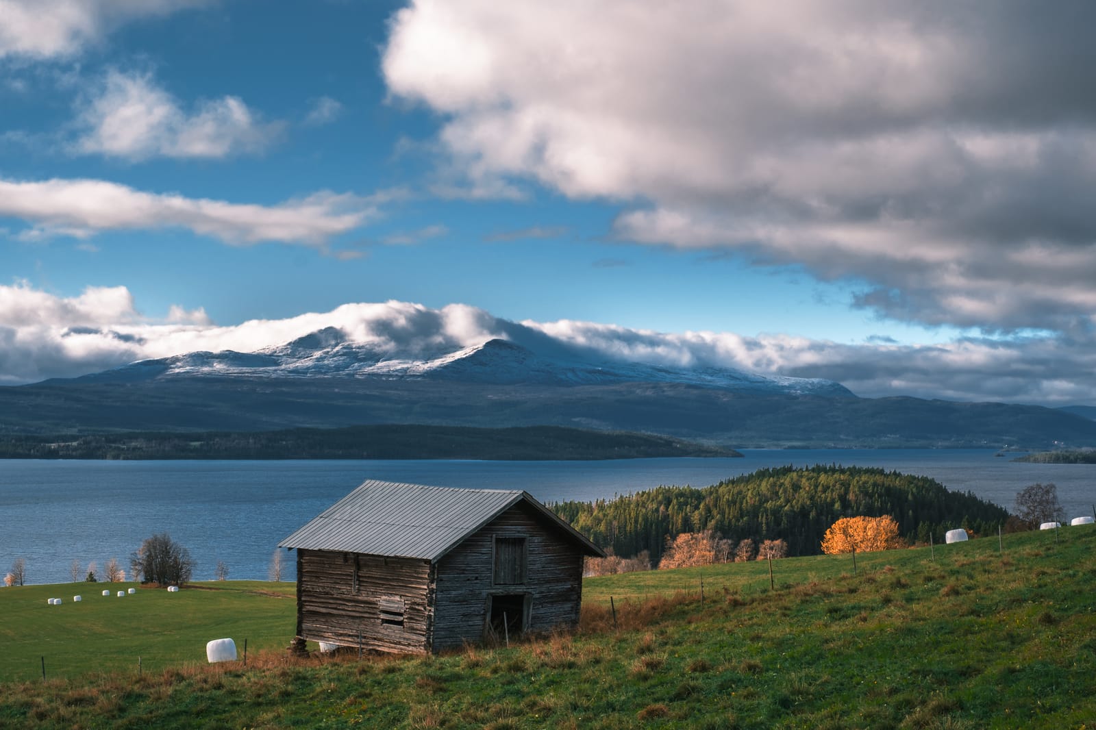 Cabaña de troncos sobre un prado junto a un lago, con montañas nevadas al fondo